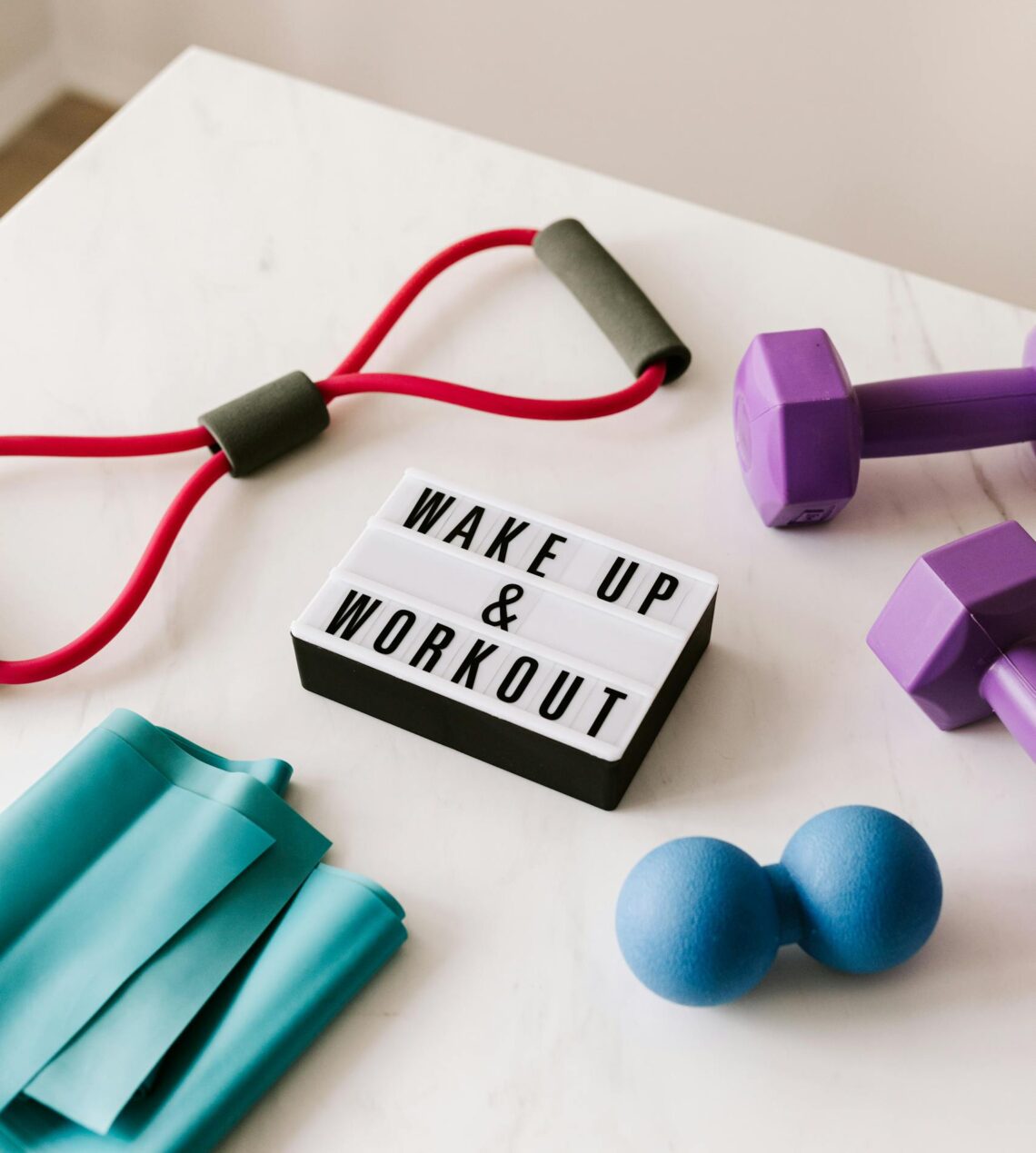 From above composition of dumbbells and massage double ball and tape and tubular expanders surrounding light box with wake up and workout words placed on white surface of table