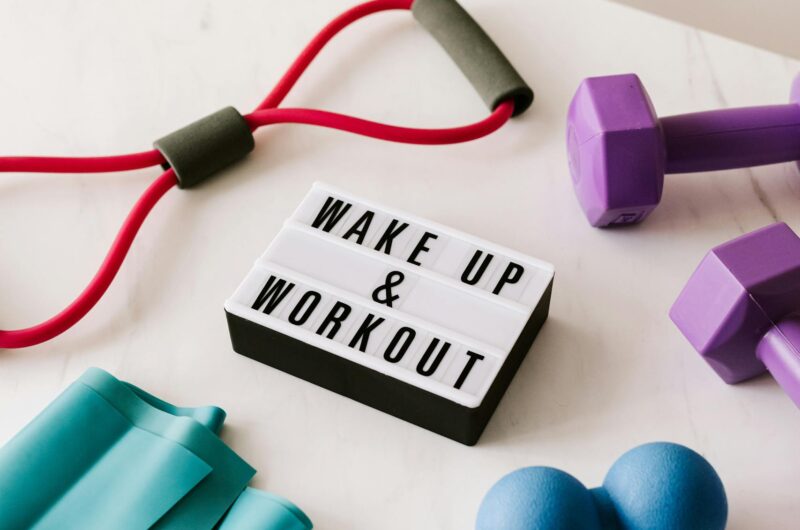 From above composition of dumbbells and massage double ball and tape and tubular expanders surrounding light box with wake up and workout words placed on white surface of table